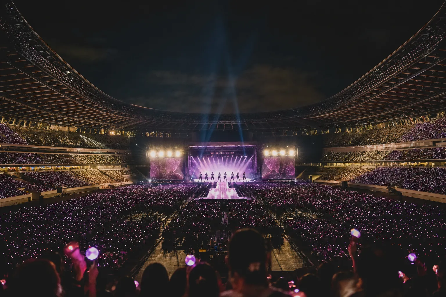 Distant K-pop group silhouettes performing at a packed Tokyo National Stadium under cinematic concert lights