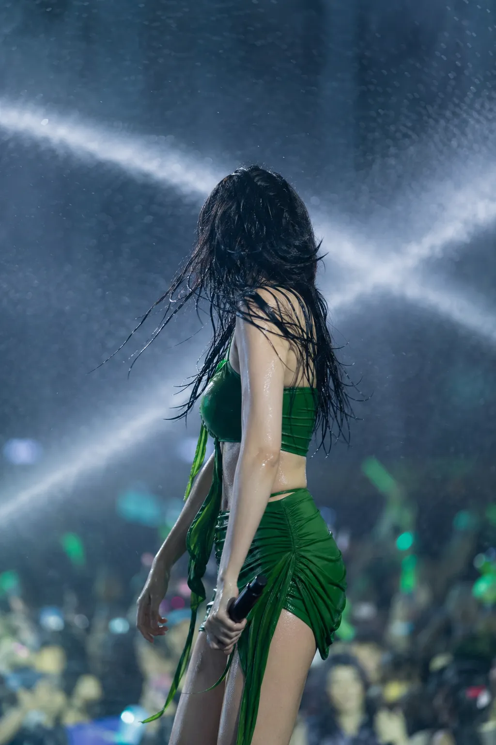 Female K-pop performer seen from behind on a wet Seoul festival stage with green lighting and water cannon effects.