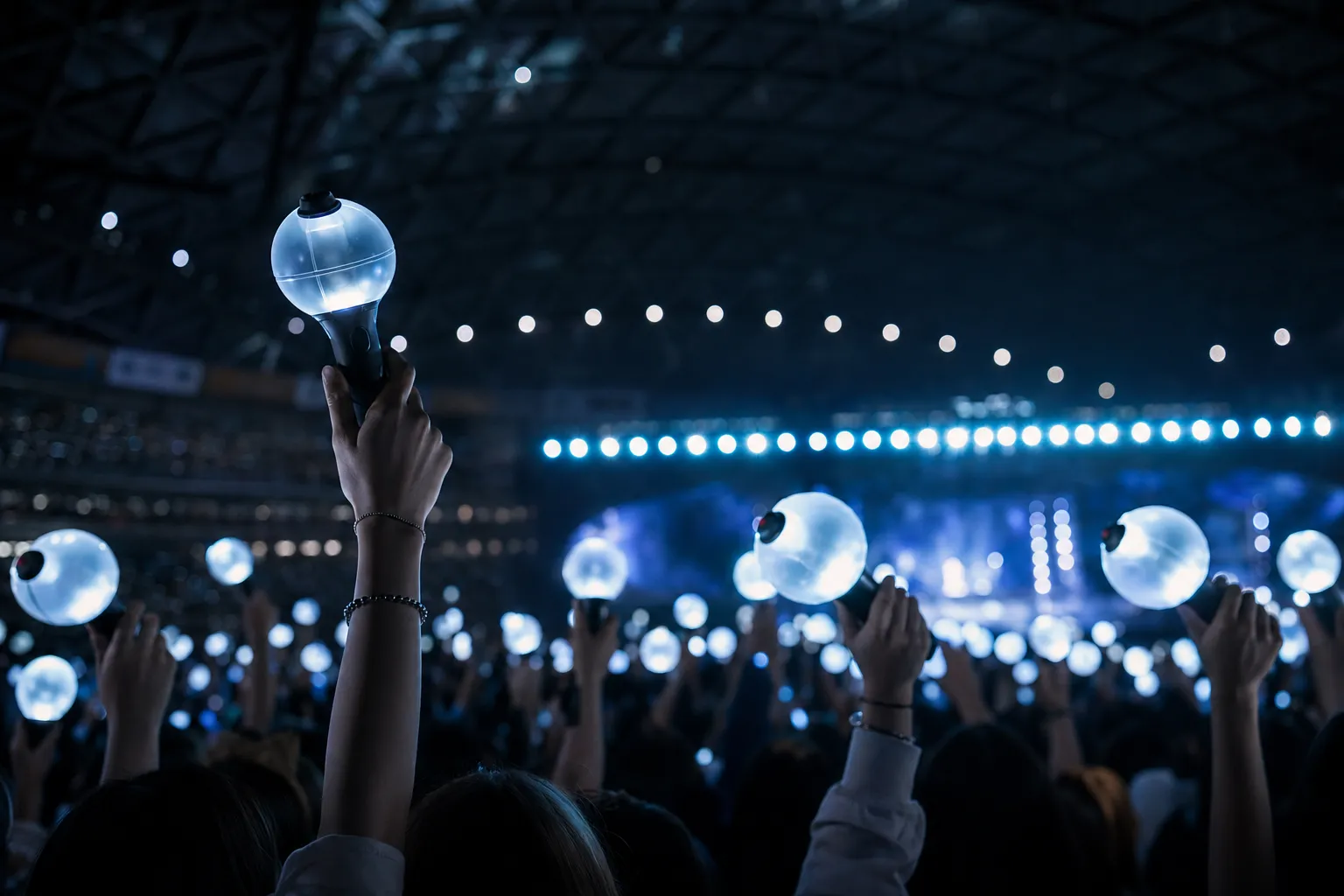Fans hold glowing lightsticks in a dome venue with blurred stage lights behind them.