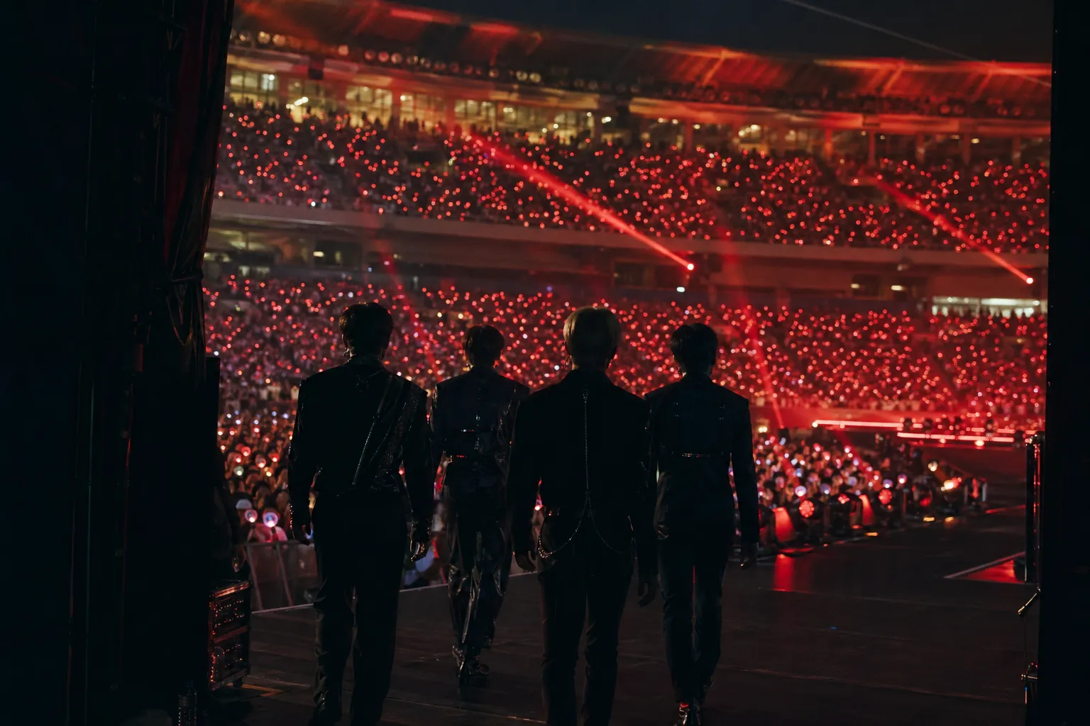 Unidentified performers face a vast red-lit stadium audience from the concert stage.