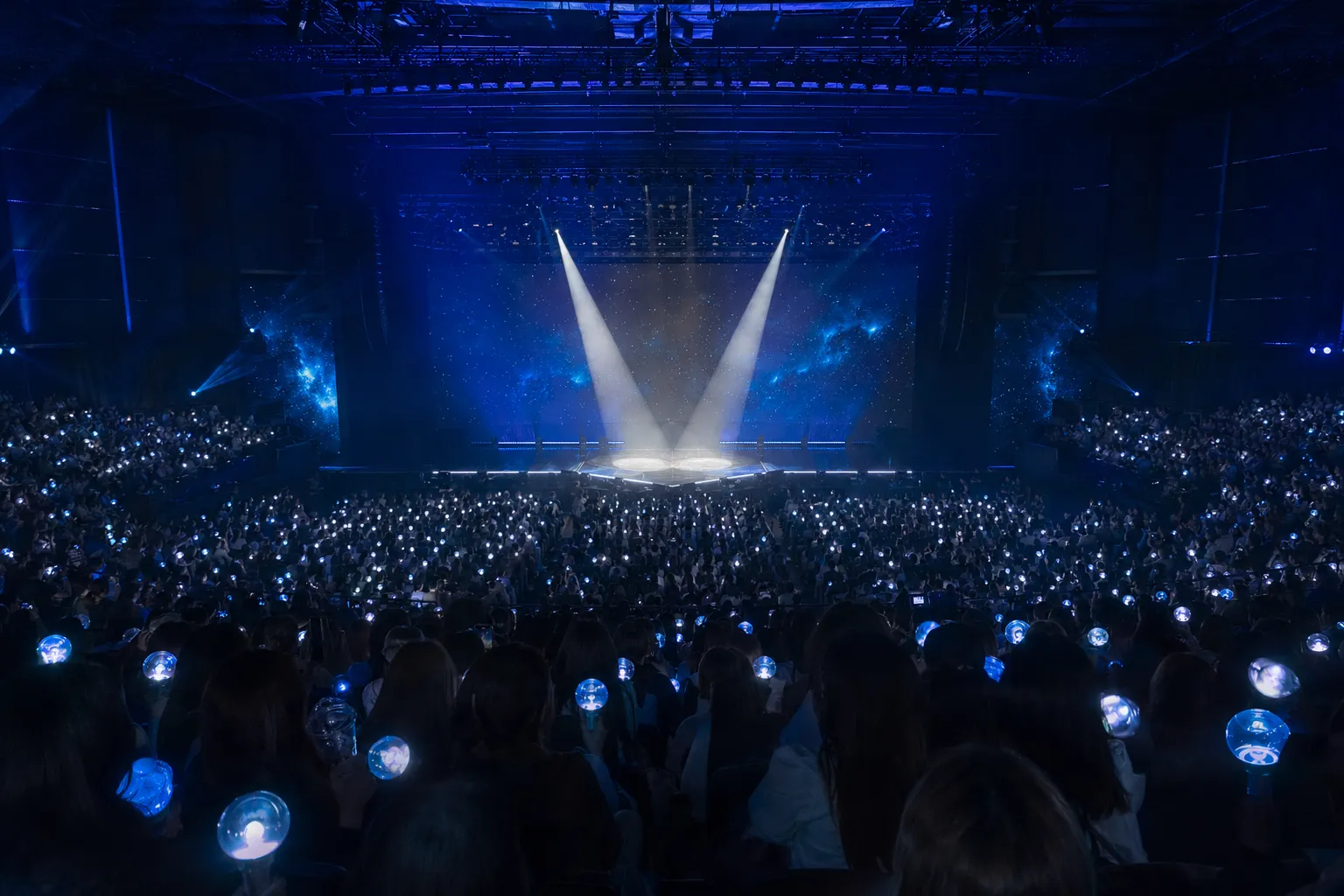 Packed K-pop fancon audience with blue light sticks facing a duo stage setup.