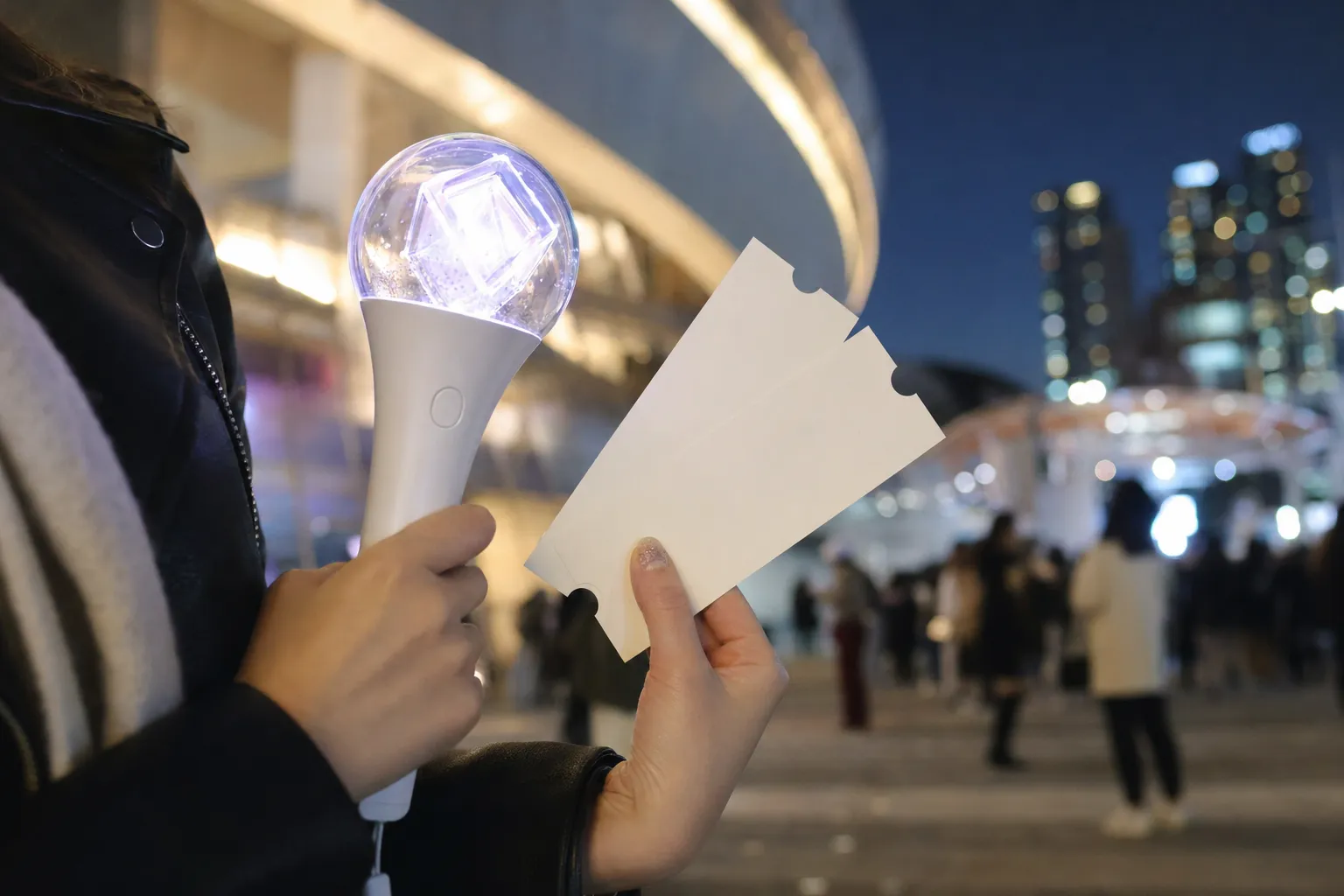 Fan holding blank concert tickets and a light stick outside a Seoul concert venue at night.